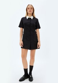 Vestido corto negro con cuello blanco de solapa, mangas cortas, dos bolsillos frontales y cierre de botones en la parte delantera. Detalle de tela texturizada.