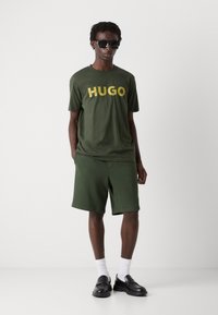 Grøn bomulds t-shirt med stort gult "HUGO" logo, parret med matchende grønne shorts og sorte sko. Accessories omfatter sorte solbriller.