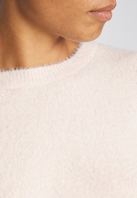 Hellrosa, weicher Strickpullover mit einem runden Ausschnitt und leicht flauschiger Textur, die ein dezentes, gleichmäßiges Muster über die gesamte Fläche zeigt.