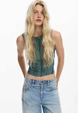 Blonde Frau trägt ärmelloses, dunkelgrünes, strukturiertes Oberteil, hellblaue Jeans und silbernen Kettengürtel mit herzförmigem Detail, vor weißem Hintergrund.