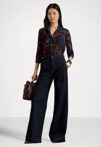 Kobieta w ciemnej bluzce w wzory i szerokich ciemnych jeansach trzyma bordową torebkę, nosi kolczyki koła i duże bransoletki na tle jednolitego tła.