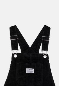 Černé kordurové salopety mají široké ramínka s kovovými sponami, vertikální žebrování a přední kapsu se značkovou etiketou.