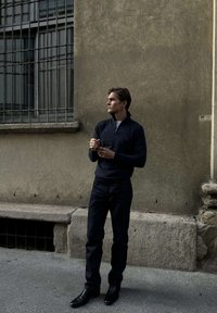 Fiatal férfi sötét ruhában áll egy városi utcán, egy texturált fal előtt, oldalra nézve, kezeit maga előtt összekulcsolva.