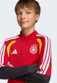 Κόκκινη αθλητική φανέλα με φερμουάρ στον γιακά, λευκές και κίτρινες λεπτομέρειες, τρεις λευκές ρίγες στα μανίκια και λογότυπο της Γερμανικής Ποδοσφαιρικής Ομοσπονδίας.