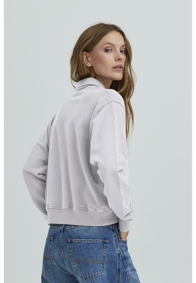 Siva zip-up dukserica s visokim ovratnikom i rebrastim manžetama, u kombinaciji s plavim trapericama. Glatka tekstura i ležeran kroj su jasno vidljivi.