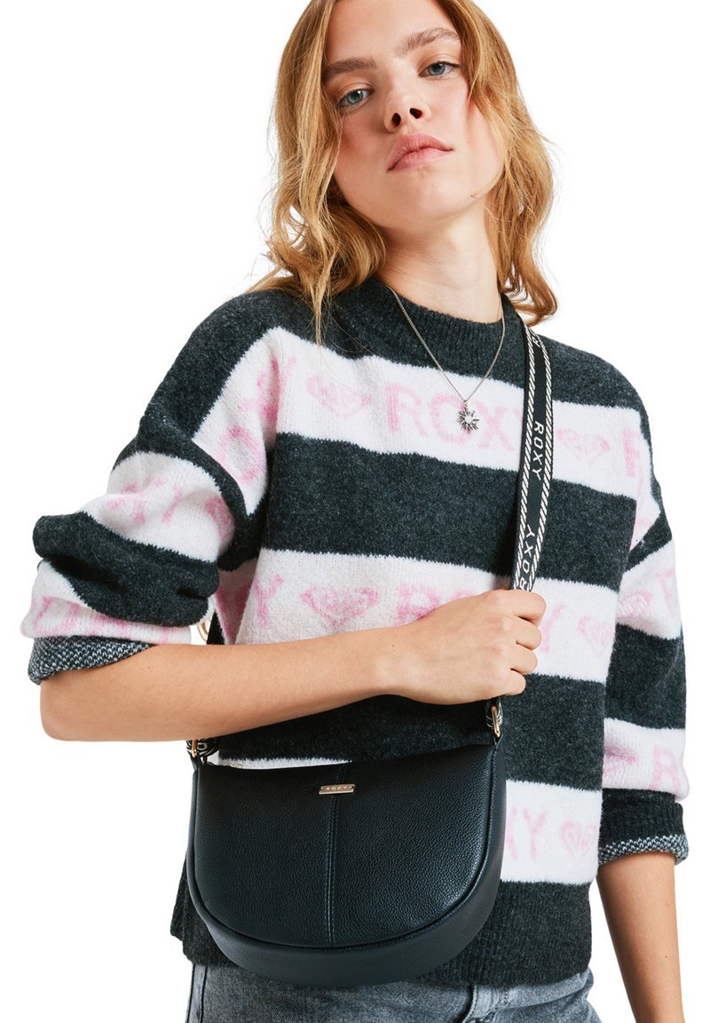 Młoda kobieta w czarno-biały sweter w paski z różowym napisem, trzymająca czarną skórzaną torbę na ramię z logo na pasku.
