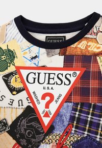 Kurzarm-Sweatshirt mit Rundhalsausschnitt, das ein Patchwork-Design aus verschiedenen Materialien, Farben und Mustern zeigt, mit einem auffälligen roten dreieckigen GUESS-Logo.