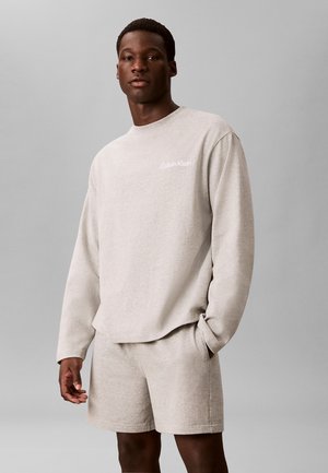 Homme portant un sweat-shirt beige clair à manches longues avec le logo "Calvin Klein" et un short assorti, debout avec une main dans la poche sur fond gris.