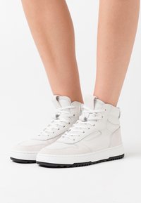 Sneakers bianchi alte con lacci indossati su gambe nude, con dettagli in suede e suole in gomma nera su uno sfondo bianco.
