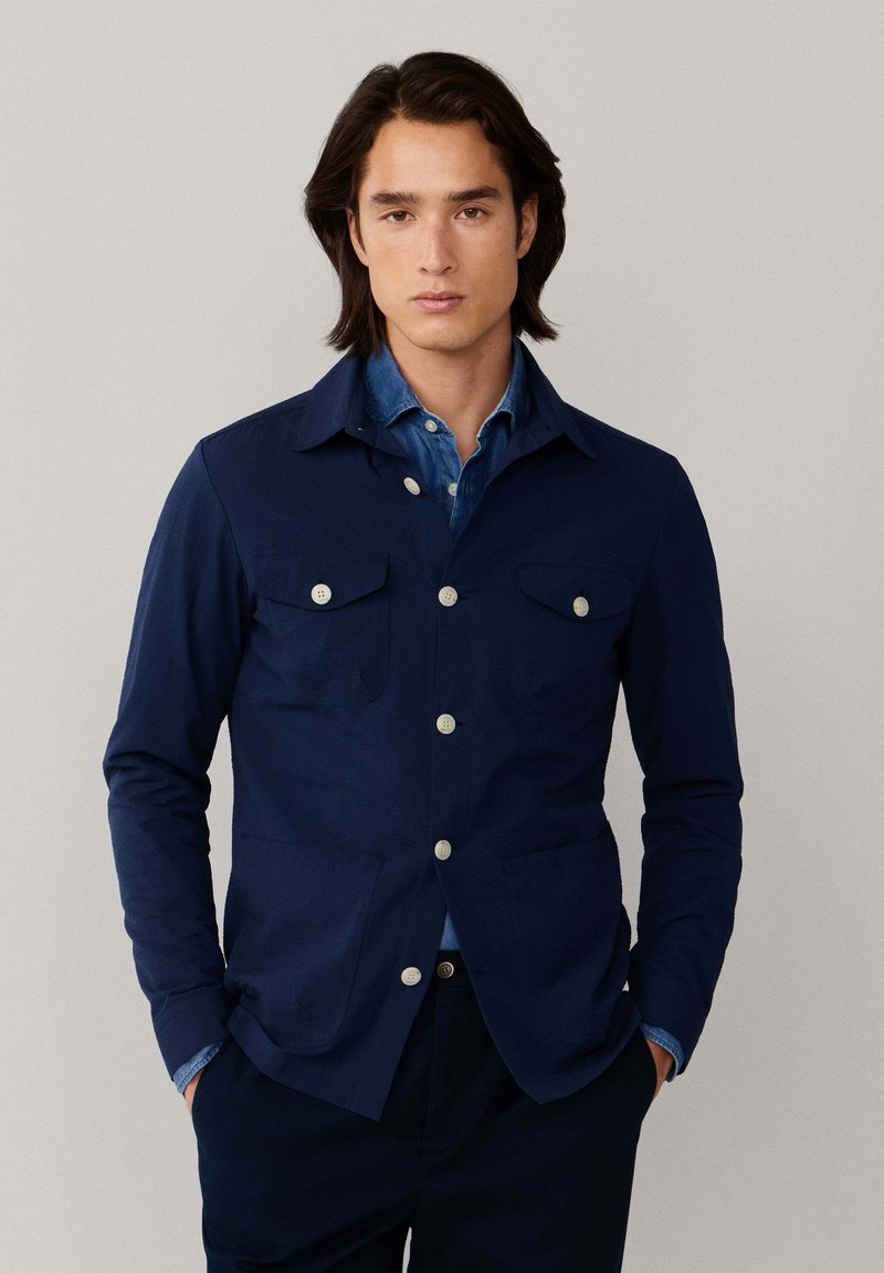 Jeune homme aux cheveux bruns mi-longs portant une veste bleu marine boutonnée sur une chemise en denim, debout devant un fond clair uni.