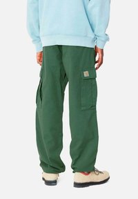 Groene cargobroek met zijvakken, ontspannen fit en zichtbare stiksels; gecombineerd met een lichtblauwe trui en beige schoeisel.