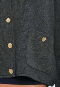 Tmavošedý sveter vyrobený z pleteného materiálu, s zlatými gombíkmi, prednou vreckom a rebrovanými detailami na leme. Detail textúrovaného materiálu zblízka.