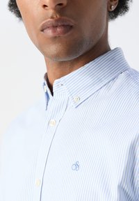 Lys blå og hvid stribet button-up skjorte med krave, featuring et lille broderet logo på brystet og hvide knapper.