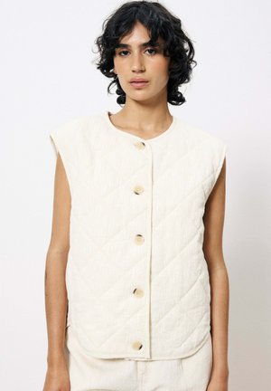 Gilet matelassé en tissu crème doux avec un col rond, des manches courtes et cinq boutons avec un mélange d'accents crème et argent.