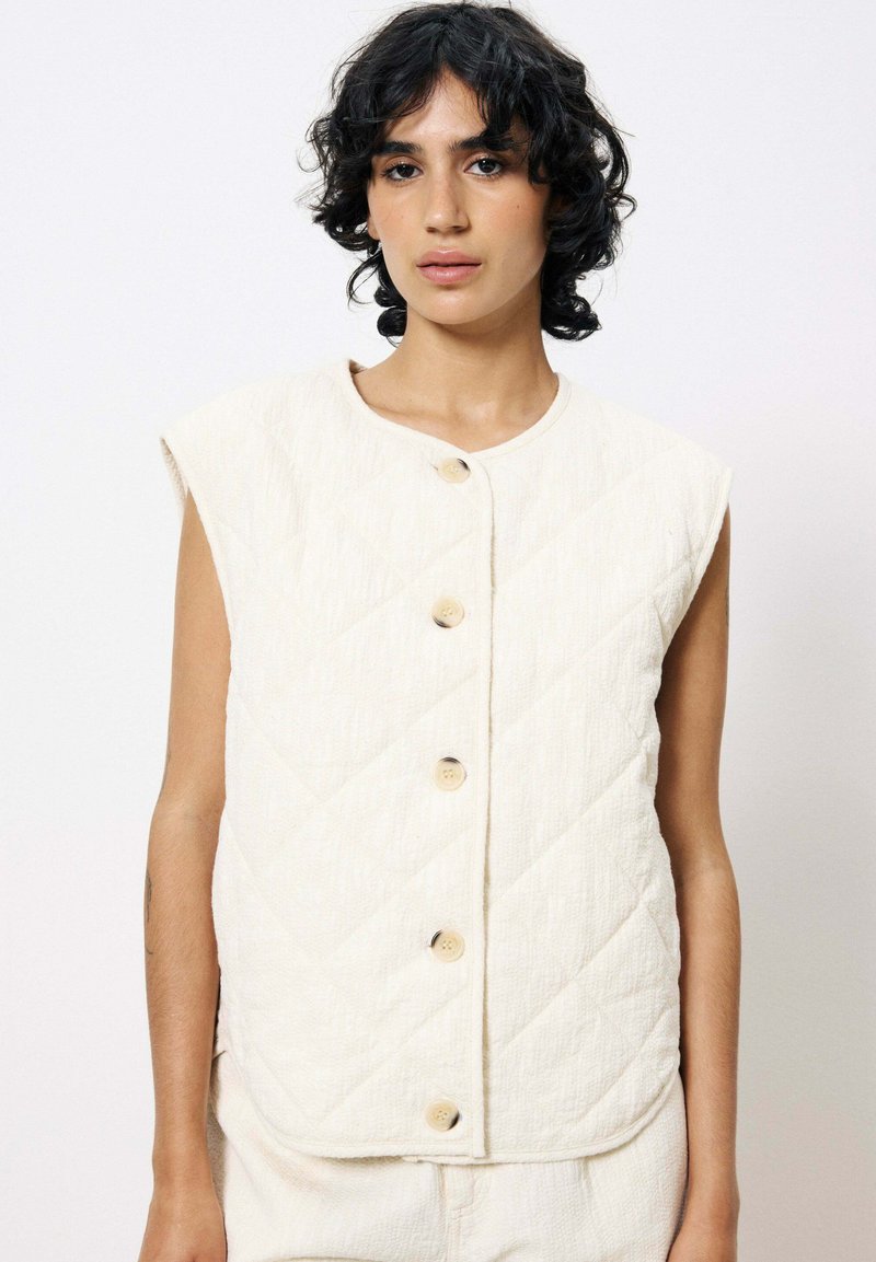 Gilet matelassé en tissu crème doux avec un col rond, des manches courtes et cinq boutons avec un mélange d'accents crème et argent.