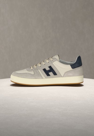 Beiger und grauer Low-Top-Sneaker aus Wildleder mit schwarzem "H"-Logo, cremefarbener Sohle und schwarzer Fersenschlaufe, platziert auf hellgrauer Oberfläche.