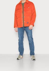 Hombre vestido con una chaqueta naranja de botones sobre una camisa verde, jeans azules y zapatillas beige, frente a un fondo blanco liso.