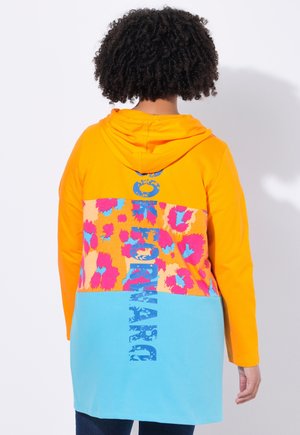 Un sweat à capuche avec un haut orange, un motif floral rose et bleu au milieu, et une partie inférieure turquoise comportant du texte.