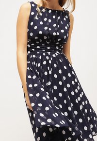 Marineblaue Polka-Dot-Kleid, ärmellos mit tailliertem Oberteil und fließendem Rock. Der Stoff ist leicht und hat große weiße Punkte sowie eine glatte Textur.