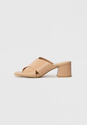 Beige åpen tå mule sandal med kryssede skinnstropper og blokkhelt på hvit bakgrunn.