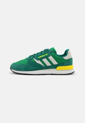 Zielony sneaker Adidas z białymi paskami, żółtym detalem na pięcie, białą podeszwą i żółtym logo Adidas, widziany z boku na jednolitym tle.