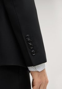 Ärmel eines schwarzen Anzugsakts mit strukturiertem Stoff, versehen mit vier schwarzen Knöpfen, und ein gemustertes weißes Hemdärmelfutter ist darunter sichtbar.