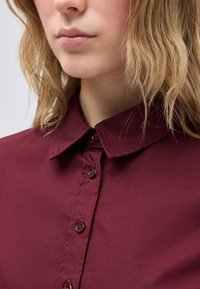 Κοντινό πλάνο ενός ατόμου που φοράει πουκάμισο με γιακά μπορντό και κουμπωμένο, με ξανθά κυματιστά μαλλιά να είναι ορατά πάνω σε φωτεινό φόντο.