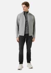 Hellgraue Jacke mit Reißverschluss über einem dunkelgrauen Oberteil, kombiniert mit schwarzen Jeans und weißen Turnschuhen. Die Jacke zeichnet sich durch ein modernes Design und eine weiche Textur aus.