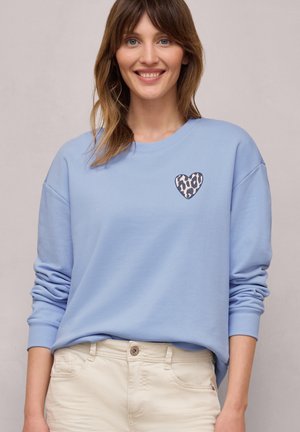 Frau lächelt, trägt hellblauen Sweatshirt mit Leopardenmuster-Herz auf der Brust und cremefarbene Hose, steht vor einfachem Hintergrund.