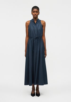 Kvinde stående i en ærmeløs mørkeblå maxikjole med bindebånd i taljen og sorte spidse hæle foran en ensfarvet hvid baggrund.