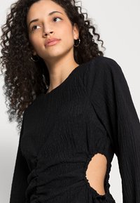 Vestido preto texturizado com mangas longas, apresentando franzido e um recorte lateral. O tecido tem uma aparência suave e amassada.