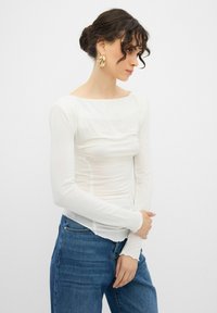 Vit långärmad topp med båturringning och rynkat tygdetalj; bärs med blå jeans med strukturerad finish och figurnära design.