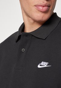 Fekete póló, texturált anyagból készült, gallérral, két gombbal és a fehér Nike logóval a bal mellkas területén.
