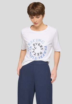 Woman with short brown hair wearing a white T-shirt with circular text and dark blue wide-leg pants, looking downward.