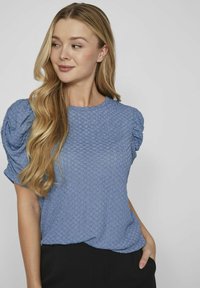 Modrá textúrovaná blúzka s krátkymi puff rukávmi, okrúhlym výstrihom a voľným strihom. Materiál má vzor včelích plástov, ktorý ponúka jemný vzhľad.