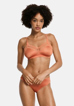 Vrouw met krullend haar, gekleed in bijpassende koraalroze satijnen beha en slip met kanten rand, staand tegen een witte achtergrond.