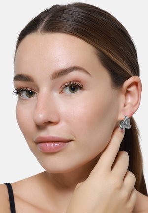 Orecchini astratti in argento con un design curvo e texture. Primo piano di un modello che si tiene l'orecchio, evidenziando i gioielli in modo prominente.