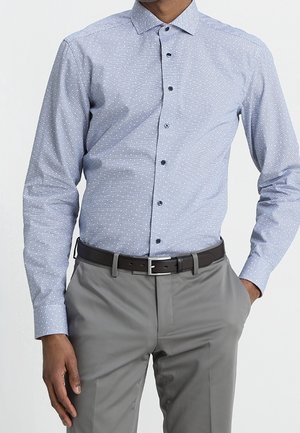 Homme portant une chemise boutonnée bleu clair à motifs rentrée dans un pantalon gris avec une ceinture noire, une main dans la poche, sur un fond uni.