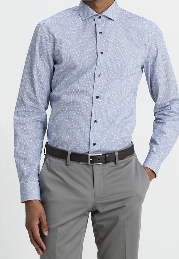 Homme portant une chemise boutonnée bleu clair à motifs rentrée dans un pantalon gris avec une ceinture noire, une main dans la poche, sur un fond uni.