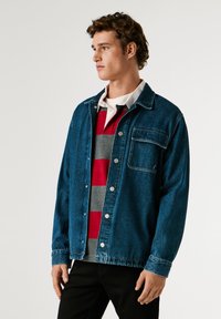 Giubbotto di denim blu scuro con chiusura a bottoni, taschino sul petto e maniche lunghe sopra una polo a righe rosse e grigie. Texture liscia.