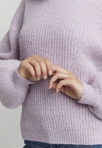 Gestrickter Pullover in hellem Lavendel, mit einem texturierten Muster, gerippten Bündchen und Saum sowie einer lockeren Passform. Die Hände sanft vorne clasped.