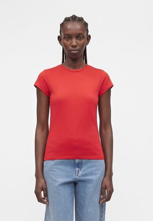 Donna con capelli intrecciati che indossa una maglietta rossa aderente a maniche corte e jeans azzurri, in piedi davanti a uno sfondo bianco semplice.