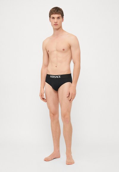 Svarte truser i brief-stil med bred midjebånd som har hvit "VERSACE"-logo. Stoffet er glatt og elastisk, noe som gir en tettsittende passform.