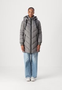 Manteau matelassé gris avec un quiltage en chevrons, fermeture éclair à l'avant, capuche avec cordons de serrage et poches latérales. Associé à un jean bleu à jambes larges et des baskets.