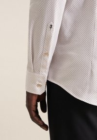 Chemise blanche à boutons avec un motif à carreaux en bleu marine et bleu clair, dotée d'un poignet avec des boutons ronds et d'un petit détail brodé.