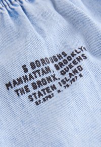 Tela azul claro con texto negro bordado que enumera los barrios de la ciudad de Nueva York y Staten Island sobre una superficie texturizada y arrugada.