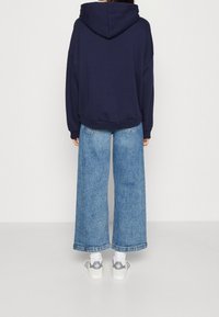 Sudadera azul marino con un diseño amplio y una capucha grande, combinada con jeans de mezclilla azul de pierna ancha y zapatos blancos con detalles en gris.