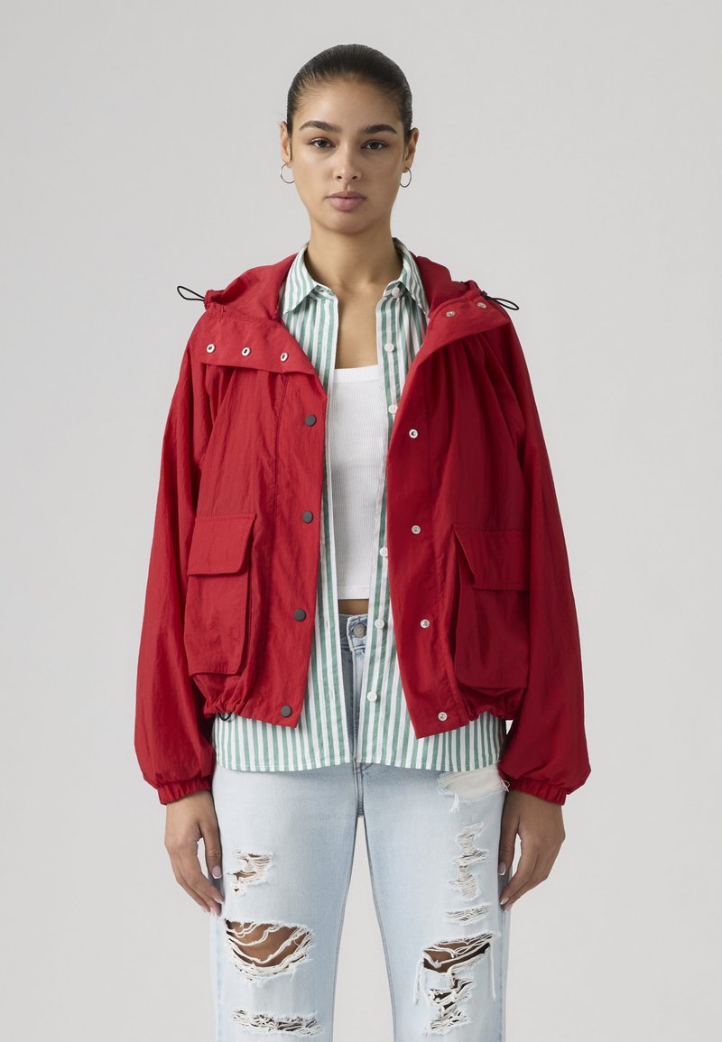 Frau trägt eine rote Jacke über einem grün-weiß gestreiften Hemd, einem weißen Crop-Top und hellblauen, abgenutzten Jeans vor neutralem Hintergrund.