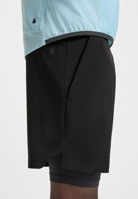 Približni prikaz osobe koja nosi crne sportske kratke hlače preko crnih kompresijskih tajica i svijetloplavu jaknu s logotipima na oba odjevna predmeta.