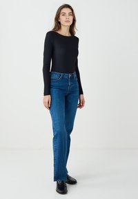 Haut noir ajusté à manches longues associé à un jean denim bleu taille haute à jambes droites. Le modèle pose devant un fond blanc uni.